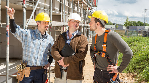 Zwei Handwerker unterhalten sich mit Steuberater vor dem Rohbau eines Hauses.