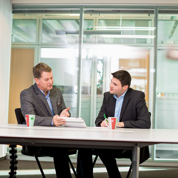 Das Bild zeigt zwei Männer, die nebeneinander sitzen und im Gespräch sind
