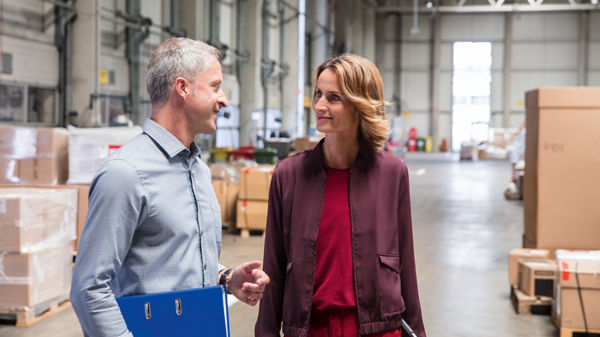 Steuerberatin und Mandant stehen in der Halle einer Logistikfirma und unterhalten sich.