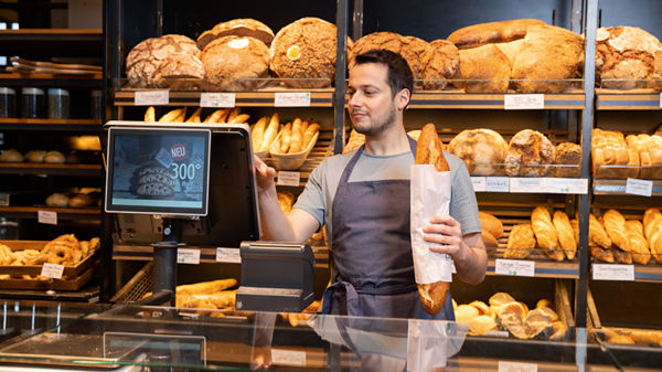 Mitarbeiter einer Bäckerei hält ein verpacktes Baguette in der Hand und gibt einen Betrag an der Kasse ein.