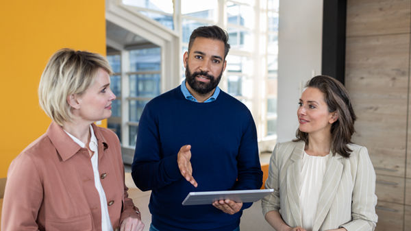 Zwei Frauen schauen auf Mann mit Tablet, der etwas erklärt.