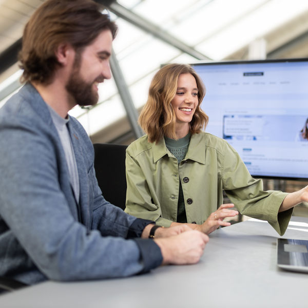 Bürosituation: Frau und Mann besprechen etwas zusammen vor Laptop, Im Hintergrund ein großer Bildschirm