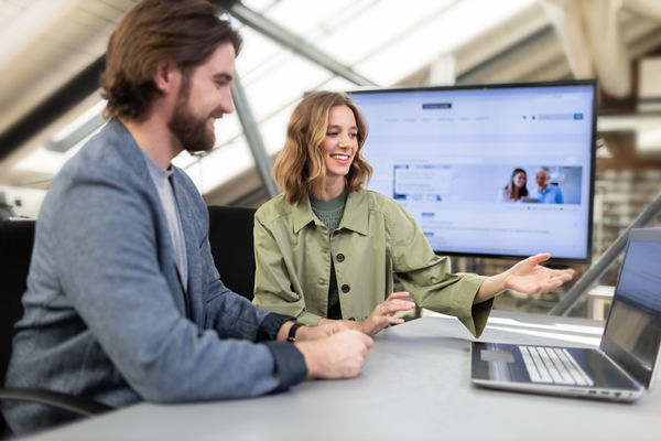 Bürosituation: Frau und Mann besprechen etwas zusammen vor Laptop, Im Hintergrund ein großer Bildschirm
