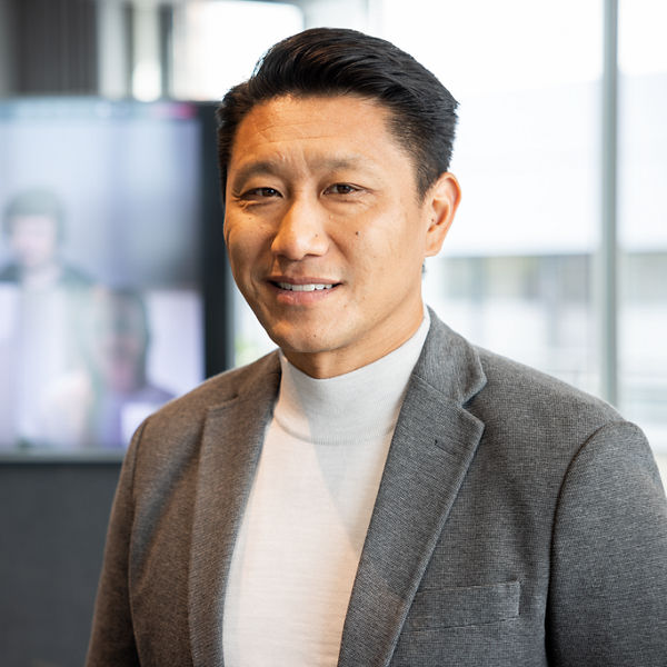 Dozent lächelt in die Kamera. Im Hintergrund ein Bildschirm mit einem virtuellen Lernraum und Lernenden (verschwommen).alt.