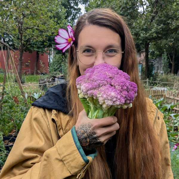 Frau mit langer brauner Mähne und Brille hält einen lilafarbenen Blumenkohl vor das Gesicht; im Haar steckt eine rosa Blüte, im Hintergrund ein Garten.