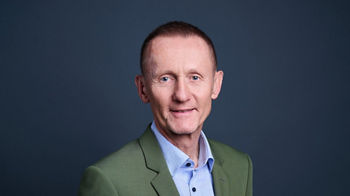 Portrait of a middle-aged man wearing a green suit and light blue dress shirt. He stands facing the camera against a dark blue studio background. The image is well-lit, highlighting his facial features and formal attire. The overall style is clean, professional, and suitable for corporate or business use.