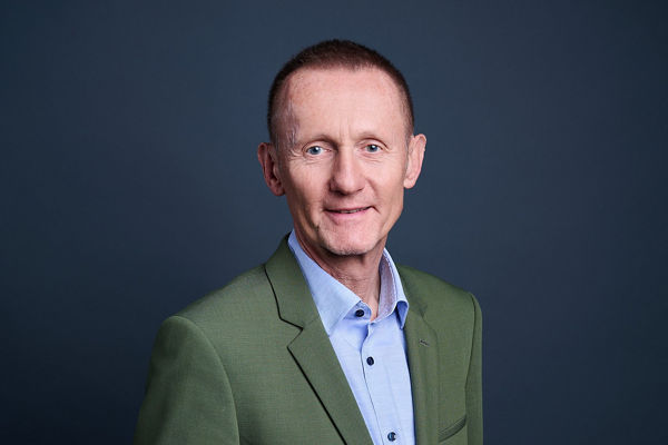 Portrait of a middle-aged man wearing a green suit and light blue dress shirt. He stands facing the camera against a dark blue studio background. The image is well-lit, highlighting his facial features and formal attire. The overall style is clean, professional, and suitable for corporate or business use.