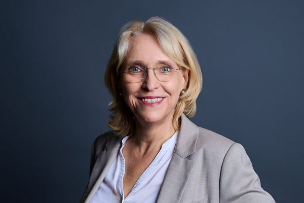 A mature woman stands confidently against a dark studio background. She wears glasses, a light blazer, and a white shirt, with one hand on her hip. The image has a professional, corporate feel suitable for business or leadership themes. Soft, even lighting highlights her friendly expression and approachable demeanor.