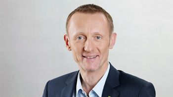 Porträt eines freundlich lächelnden Mannes im blauen Anzug mit hellblauem Hemd. Er hat kurzes hellbraunes Haar und trägt kein Zubehör. Am Revers des Sakkos ist ein kleines grünes DATEV-Logo zu sehen. Der Hintergrund ist neutral und hell.