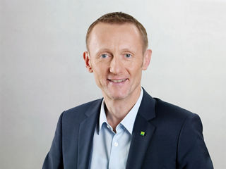 Porträt eines freundlich lächelnden Mannes im blauen Anzug mit hellblauem Hemd. Er hat kurzes hellbraunes Haar und trägt kein Zubehör. Am Revers des Sakkos ist ein kleines grünes DATEV-Logo zu sehen. Der Hintergrund ist neutral und hell.