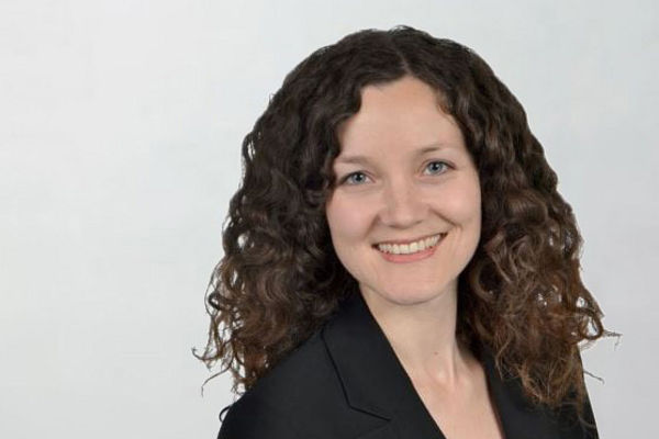 Porträt einer lächelnden Frau mit mittellangen, lockigen braunen Haaren. Sie trägt ein schwarzes Jackett über einer gemusterten Bluse. Der Hintergrund ist einfarbig und hell.