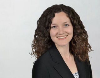 Porträt einer lächelnden Frau mit mittellangen, lockigen braunen Haaren. Sie trägt ein schwarzes Jackett über einer gemusterten Bluse. Der Hintergrund ist einfarbig und hell.