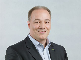 Porträt eines freundlich lächelnden Mannes mit kurzem, lichtem Haar. Er trägt ein dunkles Jackett mit dem grünen DATEV-Logo am Revers sowie ein helles Hemd. Der Hintergrund ist einfarbig und hell