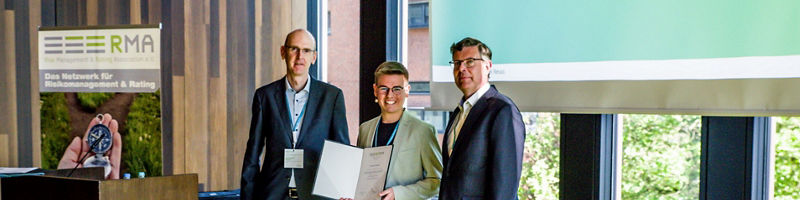 Drei Personen stehen bei einer Veranstaltung vor einem Rednerpult; eine Person hält eine Urkunde, im Hintergrund ist ein Banner des Risk Management &amp; Rating Association e. V. zu sehen.