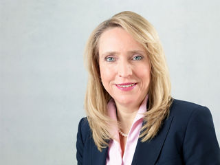 Porträt einer lächelnden Frau mit schulterlangem blondem Haar. Sie trägt ein dunkelblaues Jackett, eine rosa Bluse mit Perlenkette und dezentem Make-up. Der Hintergrund ist neutral und hell