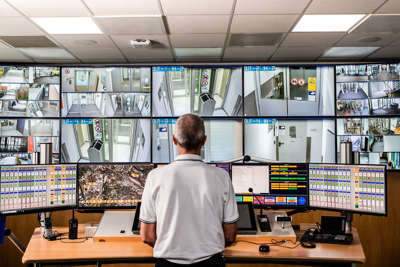 Ein Mitarbeiter in weißem Shirt steht in der DATEV-Sicherheitszentrale vor einer Wand aus Überwachungsmonitoren. Auf zahlreichen Bildschirmen sind Live-Feeds aus Kameras in Fluren, Eingangsbereichen und Außenzonen zu sehen. Auf dem Tisch sind mehrere Computer und eine Kartendarstellung eines Stadtgebiets. Das Bild vermittelt Professionalität, Überwachung und Sicherheitsmanagement.