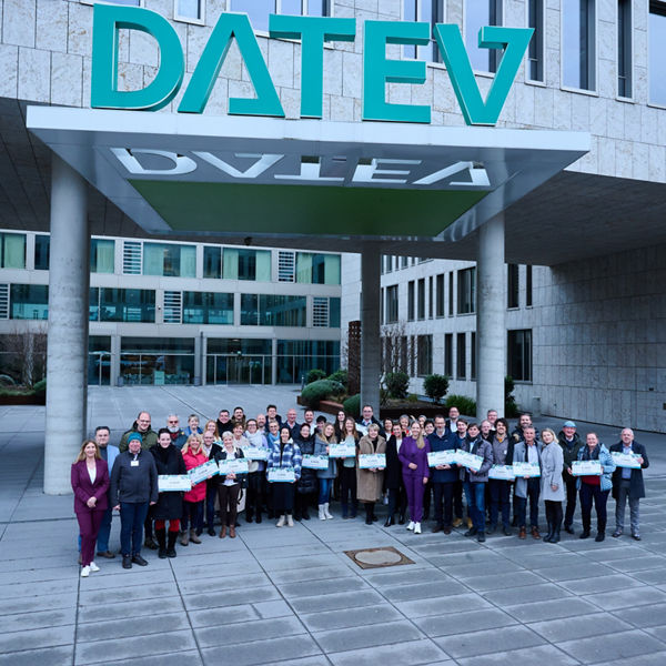 Gruppenfoto von rund 60 Personen vor dem DATEV-Hauptgebäude in Nürnberg. Die Gruppe steht unter dem großen, grünen DATEV-Schriftzug am Eingangsbereich. Viele der Teilnehmenden halten symbolische Spendenschecks in den Händen. Die Stimmung ist freundlich und gemeinschaftlich.