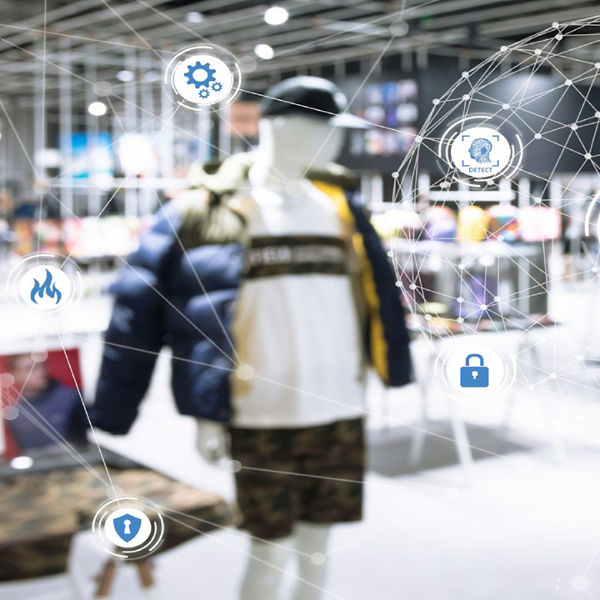 Montierte Überwachungskamera mit digital eingeblendeten Symbolen für Sicherheit, Datenschutz, Feuer- und Zugangskontrolle. Im Hintergrund ist ein moderner Einzelhandelsladen mit Bekleidung sichtbar.