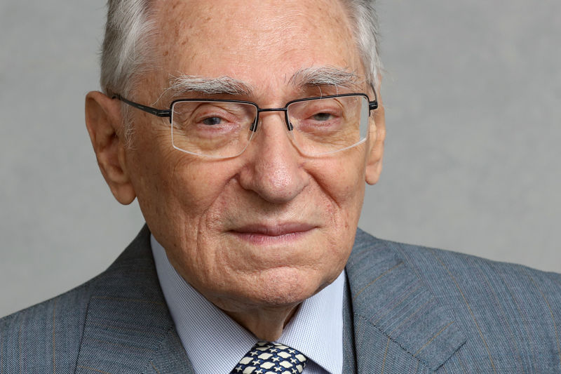 Porträt eines älteren Mannes mit grauen Haaren und Brille. Er trägt ein graues Sakko mit feinem Muster, ein helles Hemd und eine gemusterte Krawatte. Der Gesichtsausdruck ist ruhig und freundlich. Der Hintergrund ist neutral und hellgrau