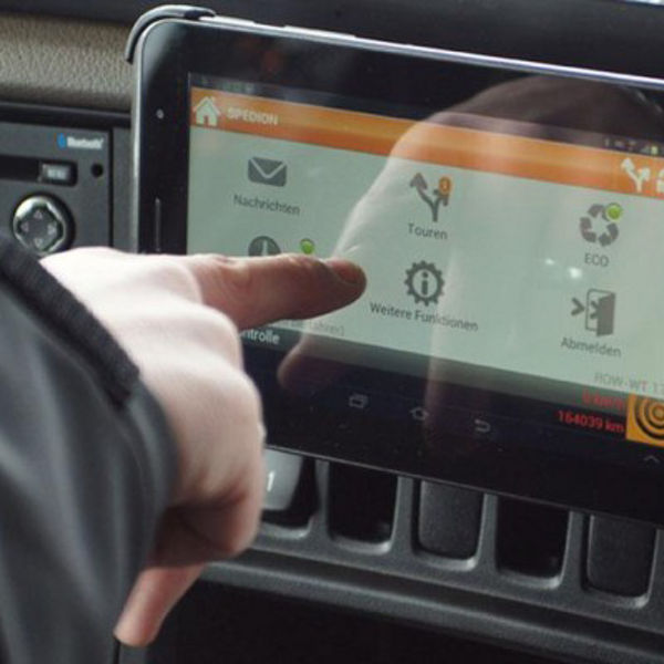 Ein Finger zeigt auf ein eingebautes Samsung-Tablet in einem Fahrzeug. Die Benutzeroberfläche zeigt Funktionen wie ‚Touren‘, ‚Nachrichten‘ und ‚Abmelden‘ – vermutlich eine Anwendung zur Logistiksteuerung..