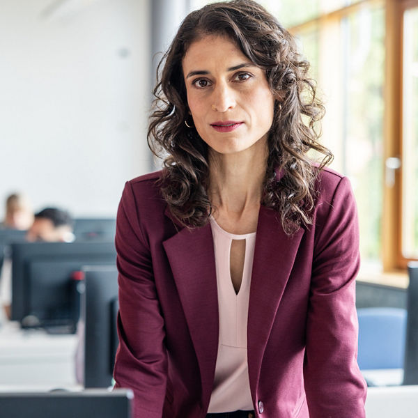 Dozentin schaut in die Kamera. Im Hintergrund ein PC-Pool mit Lernenden hinter Bildschirmen. 