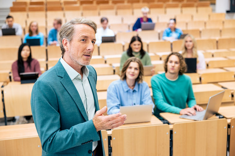 Hochschulprofessor in der Lehrsituation. Er erklärt und gestikuliert. Im Hintergrund ein Hörsaal mit Studierenden an Laptops, die aufmerksam zuhören..