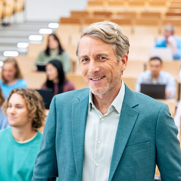 Hochschulprofessor lächelt im Hörsal in die Kamera. Im Hintergrund ein Hörsaal mit Studierenden an Laptops.
