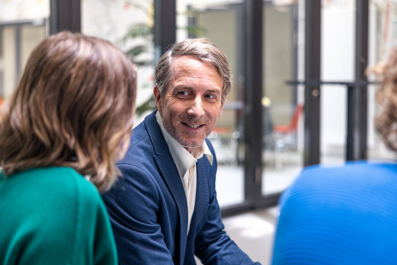 Das Bild zeigt eine Gesprächssituation zwischen einem Dozent und zwei Studierenden. Die Studierenden sitzen  mit dem Rücken zur Kamera, der Dozent schaut die links sitzende Studentin an und lächelt. 