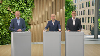 „Drei Männer in Anzügen stehen an Stehpulten in einem modernen Studio mit natürlichem Design. Im Hintergrund links ist eine begrünte Wand mit Pflanzen zu sehen, mittig eine Holzvertäfelung, rechts ein großes Fenster mit Blick auf ein modernes Bürogebäude und grüne Bäume. Alle drei schauen freundlich in die Kamera, vor ihnen stehen Wassergläser und digitale Geräte auf den Pulten.“