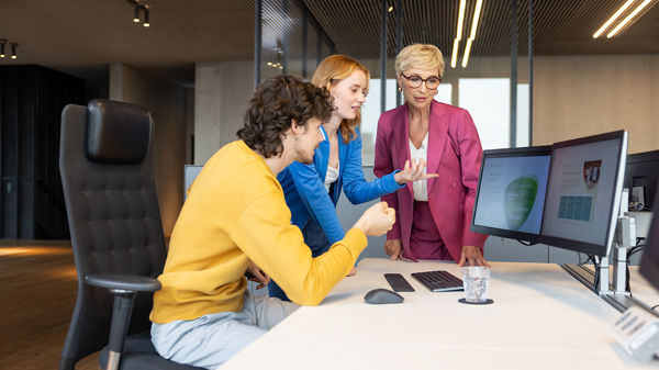 Ein junger Mann, sitzend, und zwei Damen, stehend, schauen auf einen Arbeitsplatz-Monitor. Auf dem Bild steht: Nachwuchs von morgen gewinnen und ausbilden. 