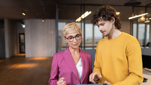 Ein junger dunkelhaariger Mann und eine mittelalte blonde Dame stehen in einer Großraumbüro-Umgebung und blicken auf ein Tablet, das der junge Mann in der Hand hält.