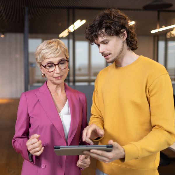 Ein junger dunkelhaariger Mann und eine mittelalte blonde Dame stehen in einer Großraumbüro-Umgebung und blicken auf ein Tablet, das der junge Mann in der Hand hält.