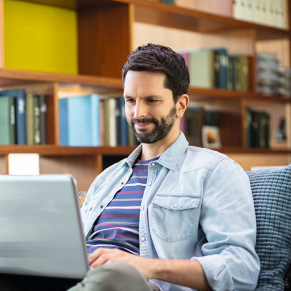 Ein junger Mann sitzt auf einem Sofa mit einem Laptop auf dem Schoß, trägt Jeansjacke und gestreiftes T-Shirt, im Hintergrund befindet sich ein Bücherregal und eine Pflanze.