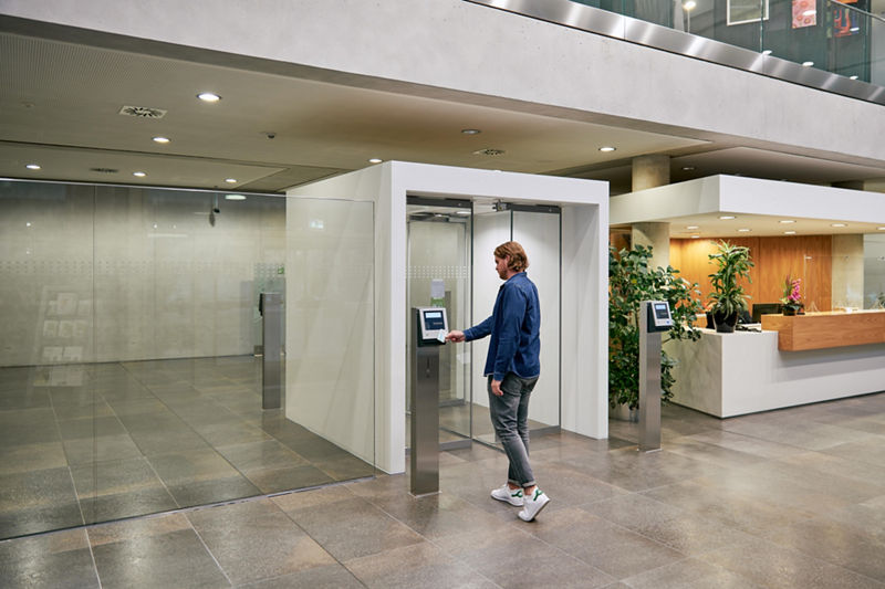 Ein Mann scannt seine Zugangskarte an einer elektronischen Zutrittskontrolle mit Drehtür in einem modernen DATEV-Gebäude. Die Umgebung ist offen gestaltet mit Glaswänden, Pflanzen und einem einladenden Empfangsbereich. Die Szene vermittelt Sicherheit, Transparenz und Besucherfreundlichkeit.