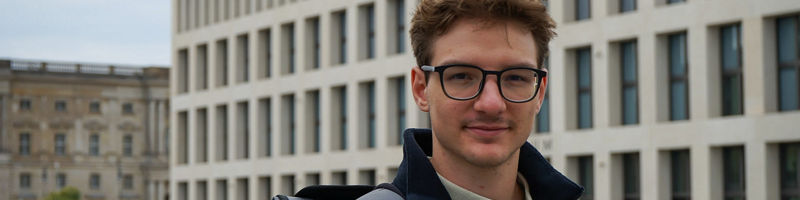 DATEV Mitarbeiter Sebastian steht mit einem Rucksack vor einem Gebäude und lächelt in die Kamera.