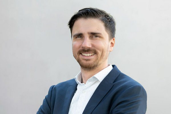 Porträt eines freundlich lächelnden Mannes mit kurzem dunklem Haar und Bart. Er trägt ein dunkelblaues Sakko über einem weißen Hemd mit feinem Muster. Die Arme sind locker verschränkt. Der Hintergrund ist neutral und hellgrau.