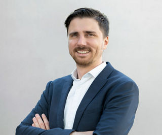 Porträt eines freundlich lächelnden Mannes mit kurzem dunklem Haar und Bart. Er trägt ein dunkelblaues Sakko über einem weißen Hemd mit feinem Muster. Die Arme sind locker verschränkt. Der Hintergrund ist neutral und hellgrau.