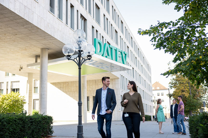 Mehrere junge Menschen gehen und unterhalten sich vor dem DATEV-Hauptgebäude in Nürnberg. Über dem Eingangsbereich prangt gut sichtbar das grüne DATEV-Logo. Im Vordergrund sind zwei Personen im Gespräch, im Hintergrund stehen weitere in kleinen Gruppen. Die Szenerie wirkt freundlich und einladend bei sonnigem Wetter.