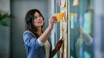 Eine Frau steht vor einer Glaswand, an der viele bunte Haftnotizen kleben. Sie trägt ein blaues Sakko und ein weißes Oberteil und befestigt gerade eine gelbe Notiz an der Wand. Die Szene wirkt konzentriert und zeigt eine kreative oder organisatorische Arbeitssituation in einem modernen Büro.