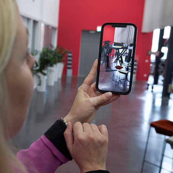 Frau fotografiert mit einem Smartphone eine Maschinenhalle mithilfe einer AR-Anwendung. Der Fokus liegt auf einem gestellten Szeneausschnitt in industrieller Umgebung mit roten Wänden und Lichtdurchflutung.