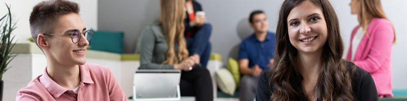 Zwei junge Erwachsene sitzen gemeinsam in einem modernen Bürobereich von DATEV und unterhalten sich.