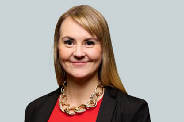 Porträt einer lächelnden Frau mit glatten, blonden, schulterlangen Haaren. Sie trägt ein rotes Oberteil, einen schwarzen Blazer und eine auffällige goldfarbene Gliederkette. Der Hintergrund ist einfarbig hellgrau.