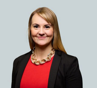 Porträt einer lächelnden Frau mit glatten, blonden, schulterlangen Haaren. Sie trägt ein rotes Oberteil, einen schwarzen Blazer und eine auffällige goldfarbene Gliederkette. Der Hintergrund ist einfarbig hellgrau.