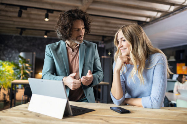 On this picture you can see two persons discussing while standing at a table with a tablet on it.