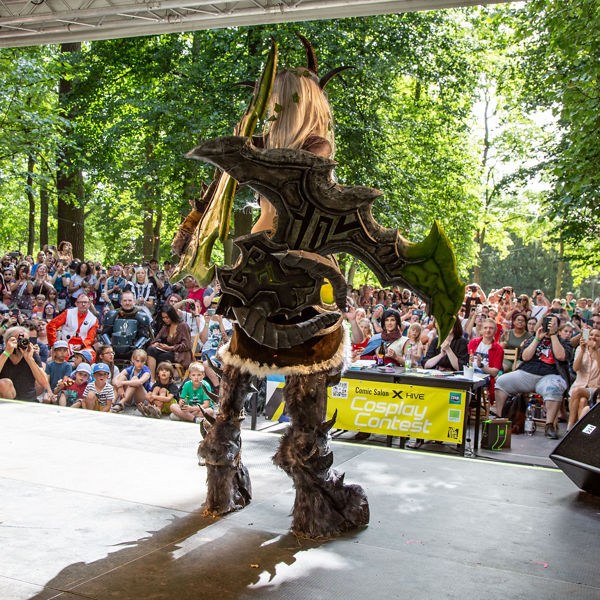 Person in aufwendigem Cosplay-Kostüm mit riesigem Schwert steht auf einer Bühne im Freien; vor der Bühne sitzt und steht ein großes Publikum, viele machen Fotos.