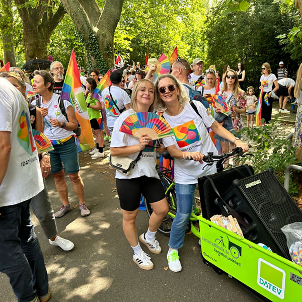 Menschen beim CSD im Park, viele tragen weiße T-Shirts mit buntem Logo und halten Regenbogenfahnen und Fächer, vor ihnen eine grüne Transportkarre mit Lautsprecher und DATEV-Logo.