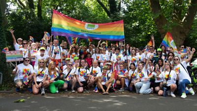 Große Gruppe von Menschen in weißen T-Shirts mit Regenbogen-Logo posiert lachend unter einem bunten DATEV-Banner mit der Aufschrift ‚#DATEVistbunt‘.