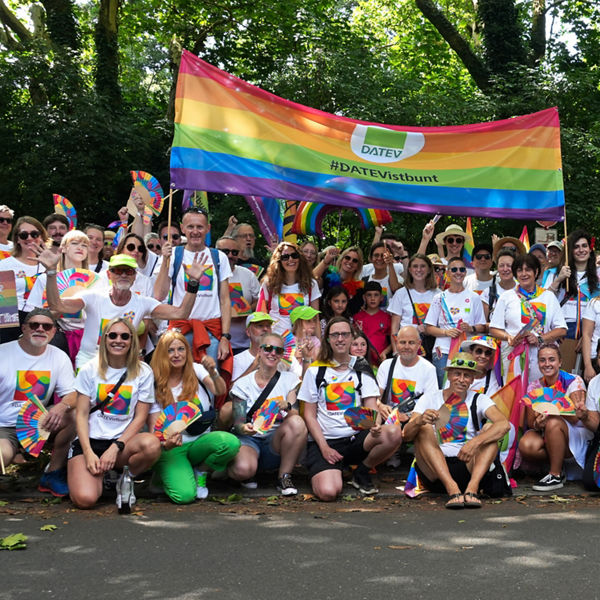 Große Gruppe von Menschen in weißen T-Shirts mit Regenbogen-Logo posiert lachend unter einem bunten DATEV-Banner mit der Aufschrift ‚#DATEVistbunt‘.