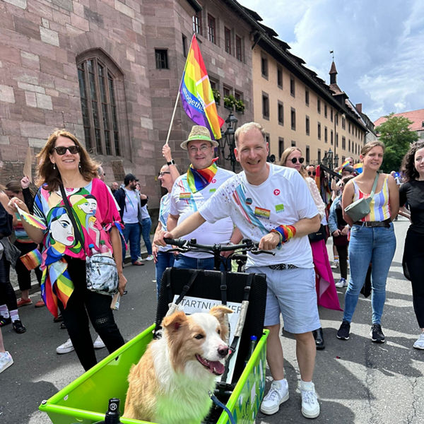 Menschen bei einer Pride-Parade, vorne ein Hund in einer grünen Lastenrad-Box; Teilnehmer tragen bunte Kleidung und Regenbogenfahnen.