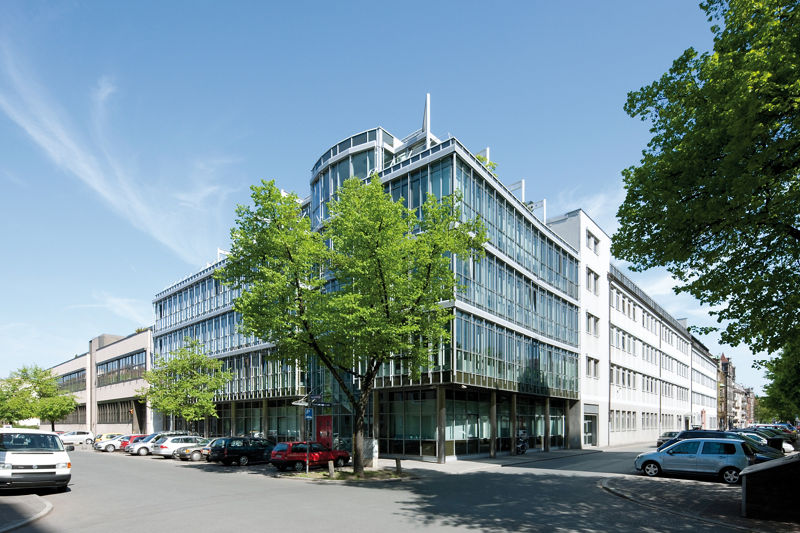DATEV-Gebäude mit Glasfassade und abgerundeter Gebäudeecke in moderner Architektur. Große Fensterflächen und markante Metallkonstruktionen prägen die Front. Vor dem Gebäude stehen junge Bäume und mehrere geparkte Autos. Eine ruhige Seitenstraße verläuft entlang des Bürokomplexes bei klarem Himmel.